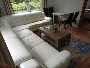 A living room with a white leather sofa, a wooden coffee table with a vase of flowers, and a dining area with a small table and chairs. - 3-room apartment on the first floor (Marienheide)