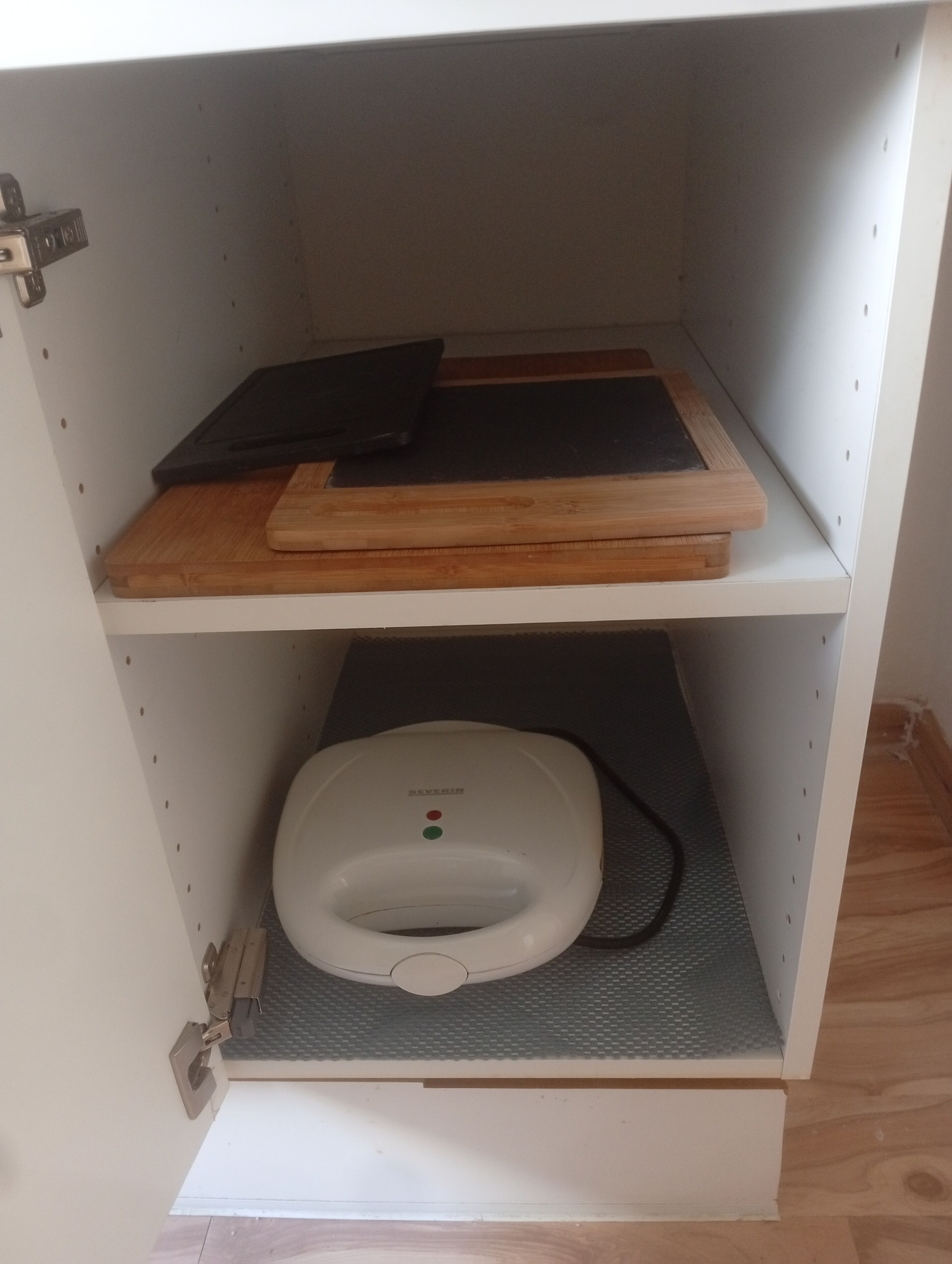 A kitchen cabinet with a cutting board, a black tray, and a white appliance.