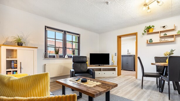 A modern living room with a sofa, a coffee table, a TV, and a dining area with chairs and a table.