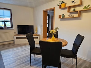 A dining area with a round table, black chairs, a yellow vase with flowers, and a television on a wooden stand.