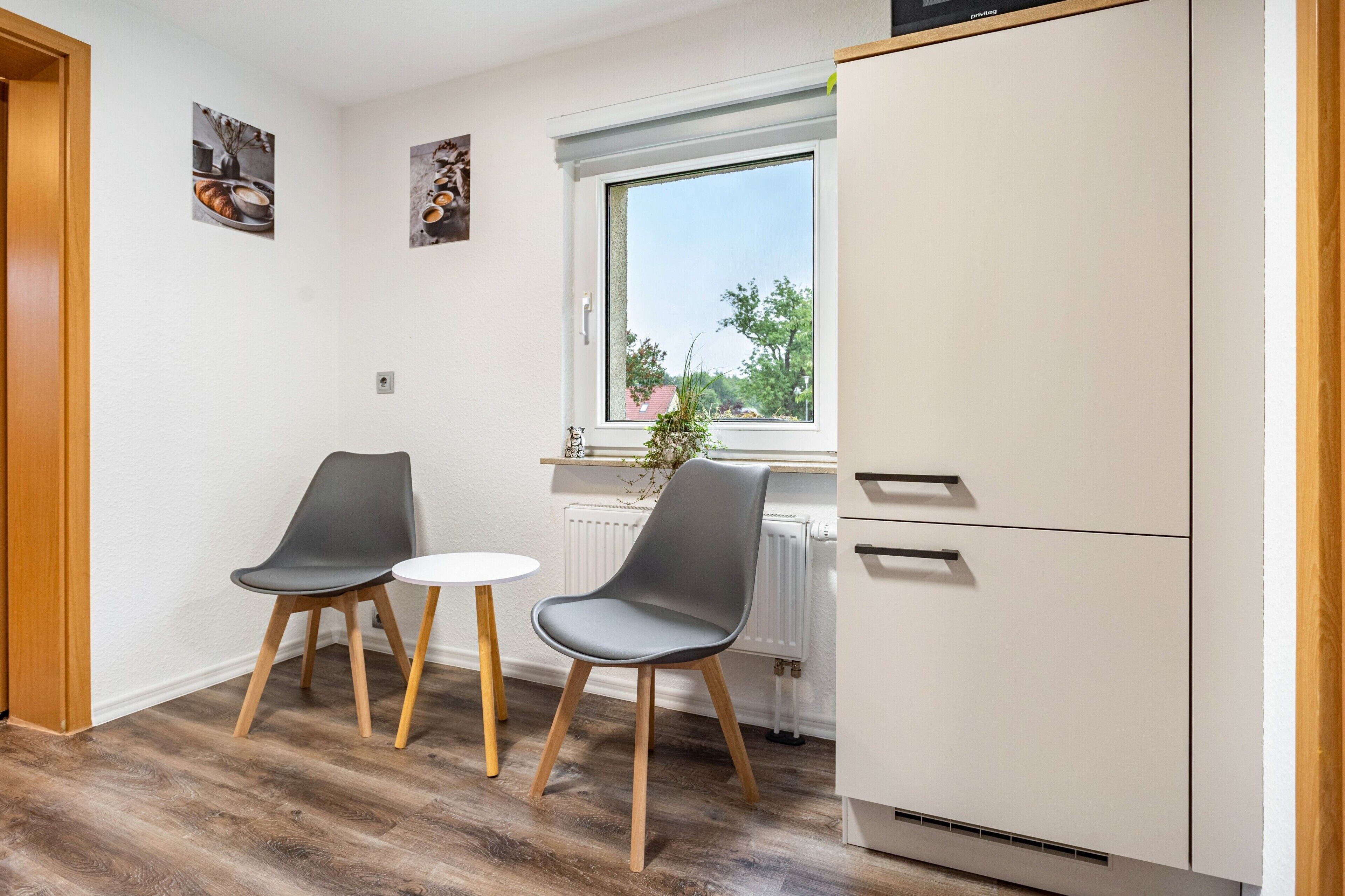 Eine kleine Küchenzeile mit einem weißen Kühlschrank, zwei grauen Stühlen, einem kleinen runden Tisch und einem Fenster mit Blick auf Grünflächen.