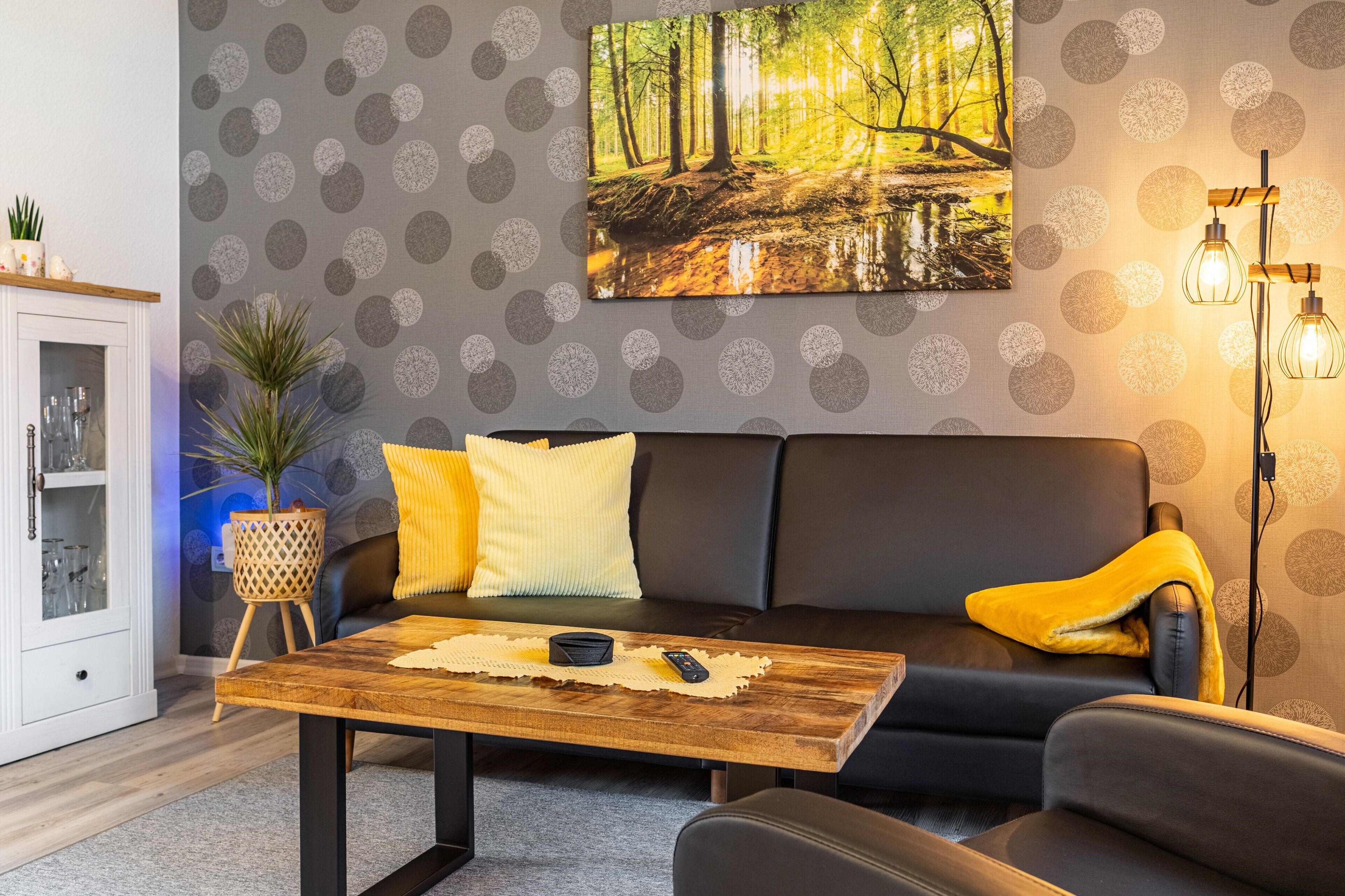 Ein modernes Wohnzimmer mit einem dunklen Sofa, einem Holztisch, einem Waldbild und einer gemusterten Tapete.