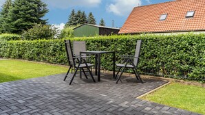 An outdoor patio with a table and chairs, surrounded by a hedge and a building with a red roof.