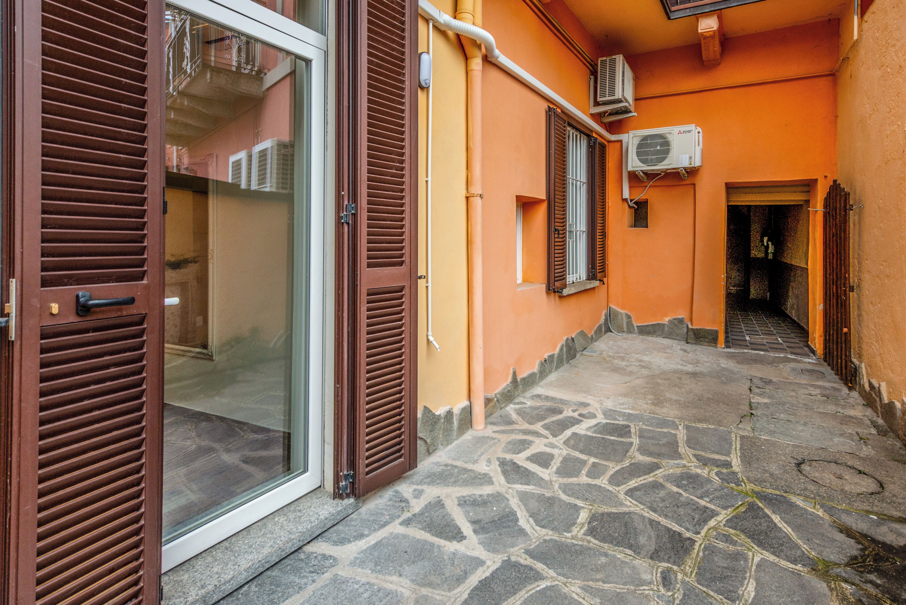 A narrow stone-paved alleyway with a glass door and brown shutters, leading to a building with orange walls and air conditioning units.