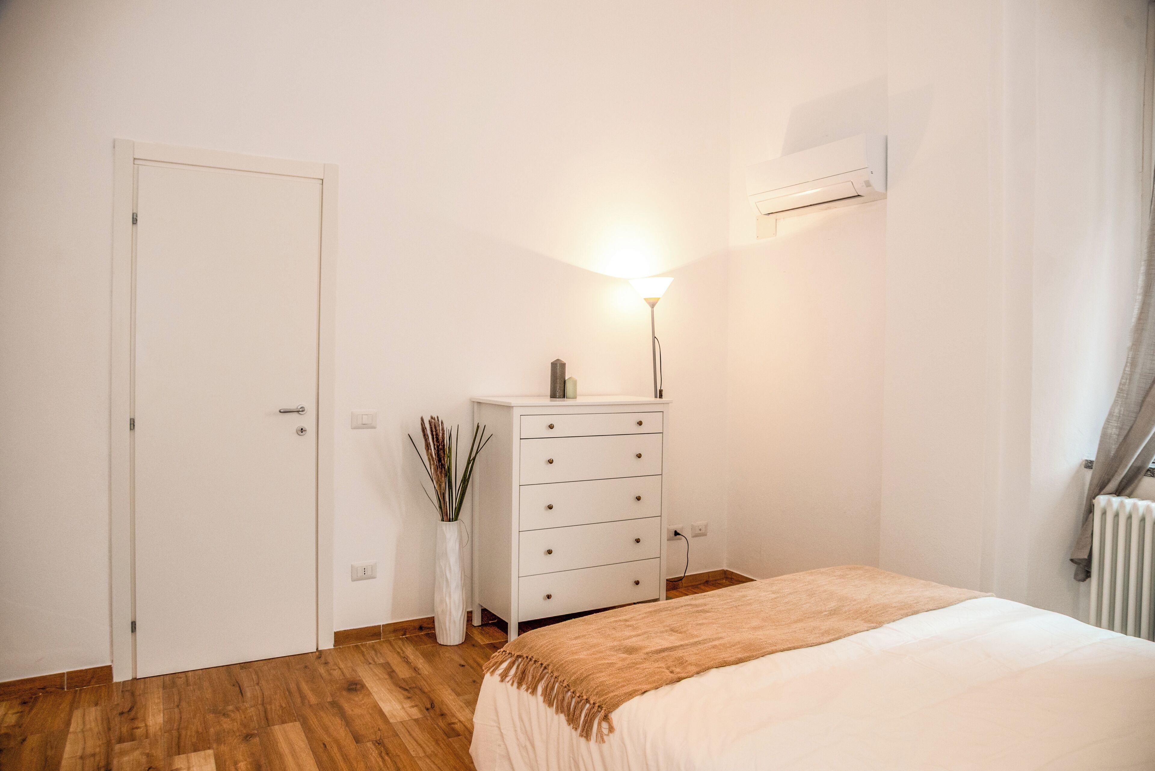 Een slaapkamer met een houten vloer, een bed met een beige deken, een witte dressoir, een lamp en een radiator.