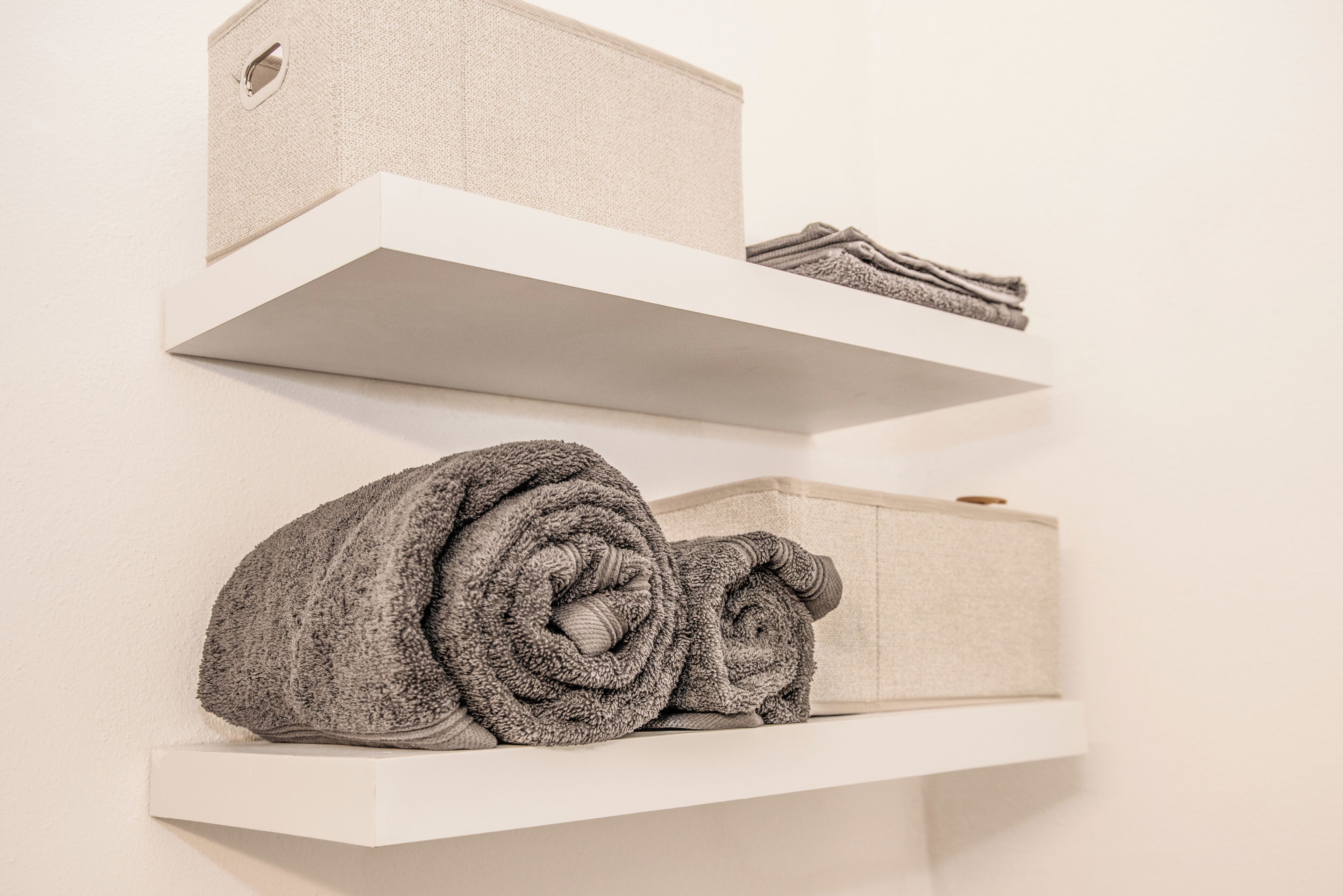 Three white shelves with folded towels and a beige box on the bottom shelf.