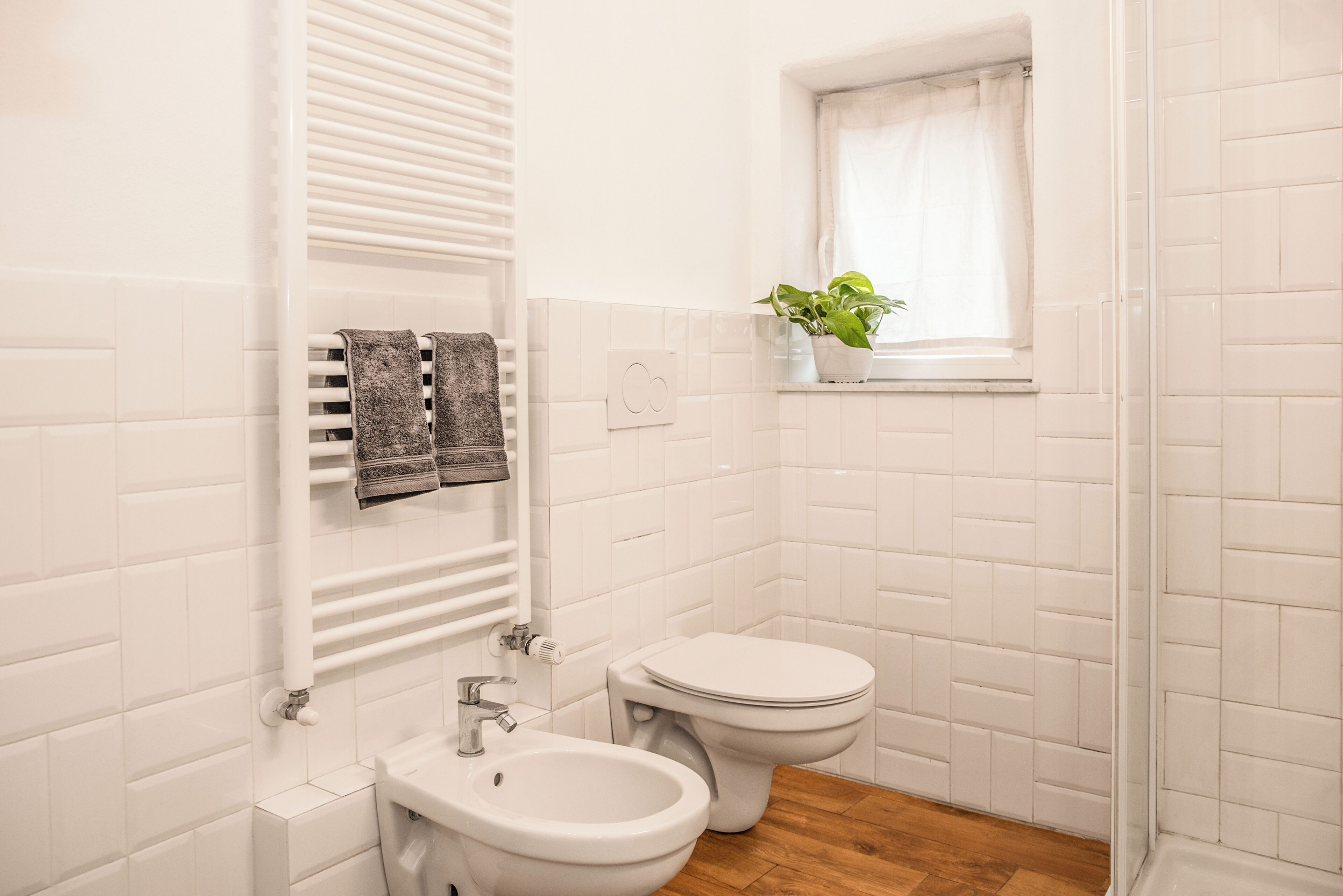 A bathroom with white tiles, a bidet, a toilet, a sink, a towel rack, and a window with a plant.