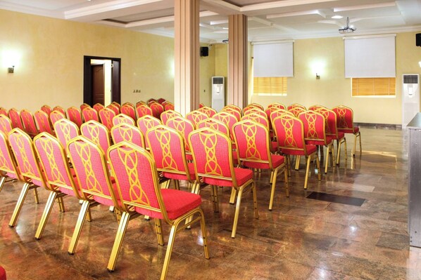 Un salón de conferencias con hileras de sillas rojas con detalles dorados, un atril y un ventilador de techo.