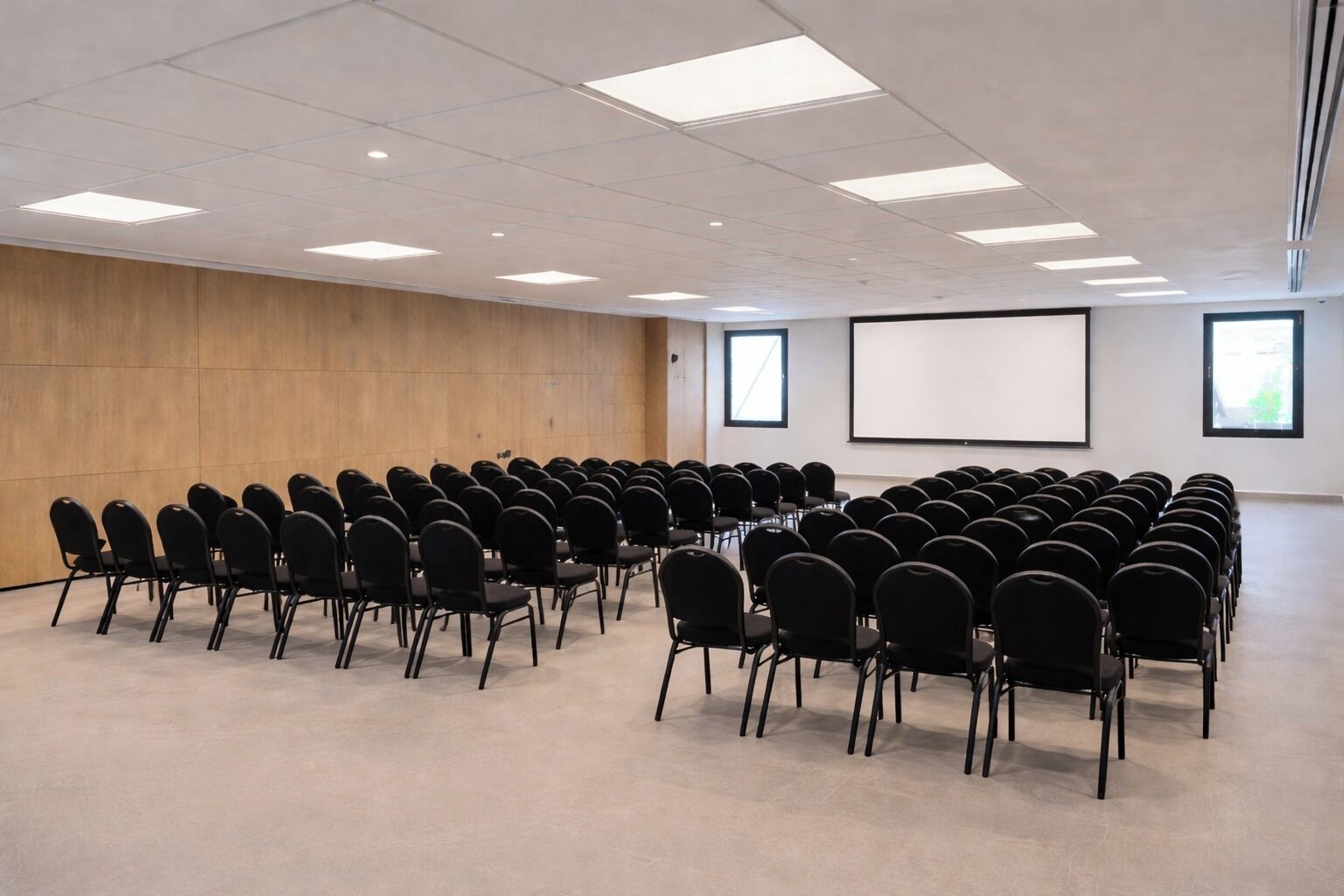 Un salón de conferencias con filas de sillas negras, una pantalla grande y una pared de madera.
