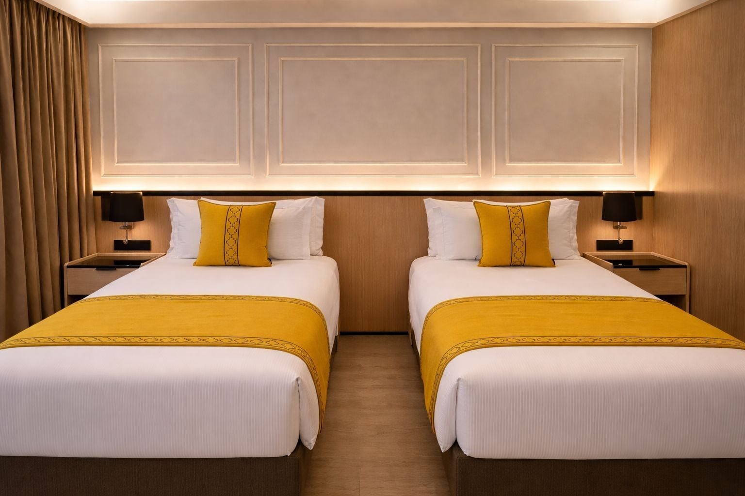 Habitación de hotel con dos camas, cada una con colcha blanca y almohada decorativa amarilla. Hay dos mesitas de noche con lámparas, y la pared es de paneles de madera.