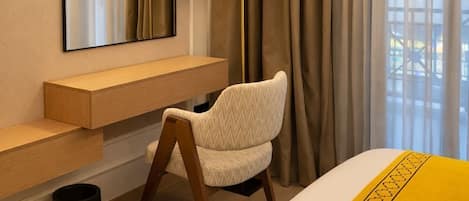 A hotel room with a bed, a chair, a desk, a mirror, a lamp, and a window with curtains.