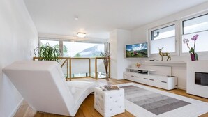 Ein modernes Wohnzimmer mit einem weißen Sofa, einem kleinen Hocker, einer weißen Unterhaltungsanlage und einem großen Fenster mit Bergblick.
