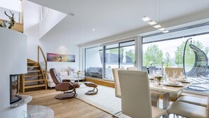 Ein modernes Wohnzimmer mit Kamin, einer Essecke mit Tisch für vier Personen und großen Fenstern mit Blick ins Freie.