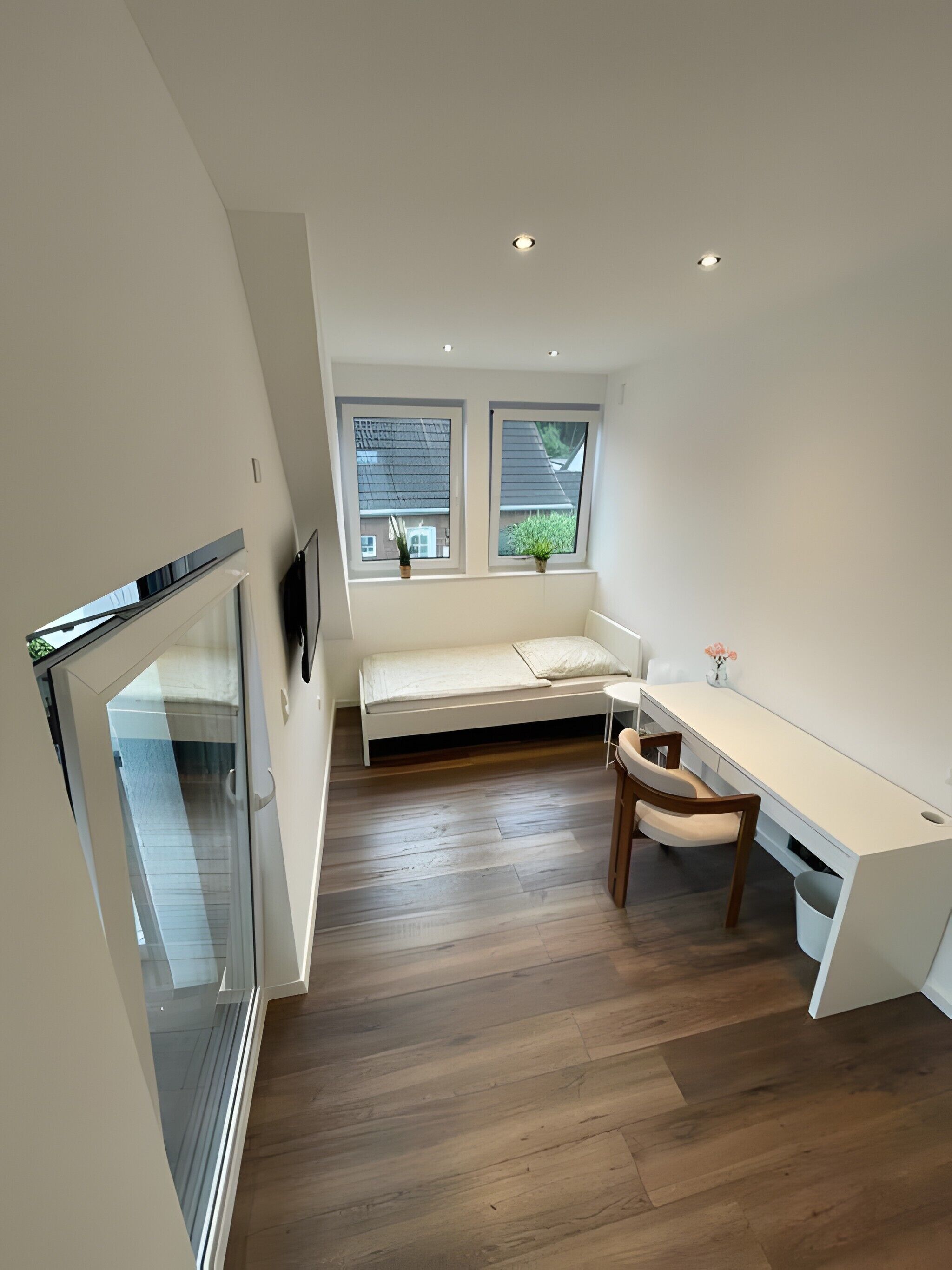 A modern, compact room with a bed, a desk, and a window with a view of a house.