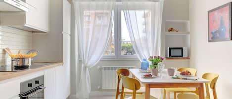 A modern kitchen with a dining area, white cabinetry, and a large window with sheer curtains.