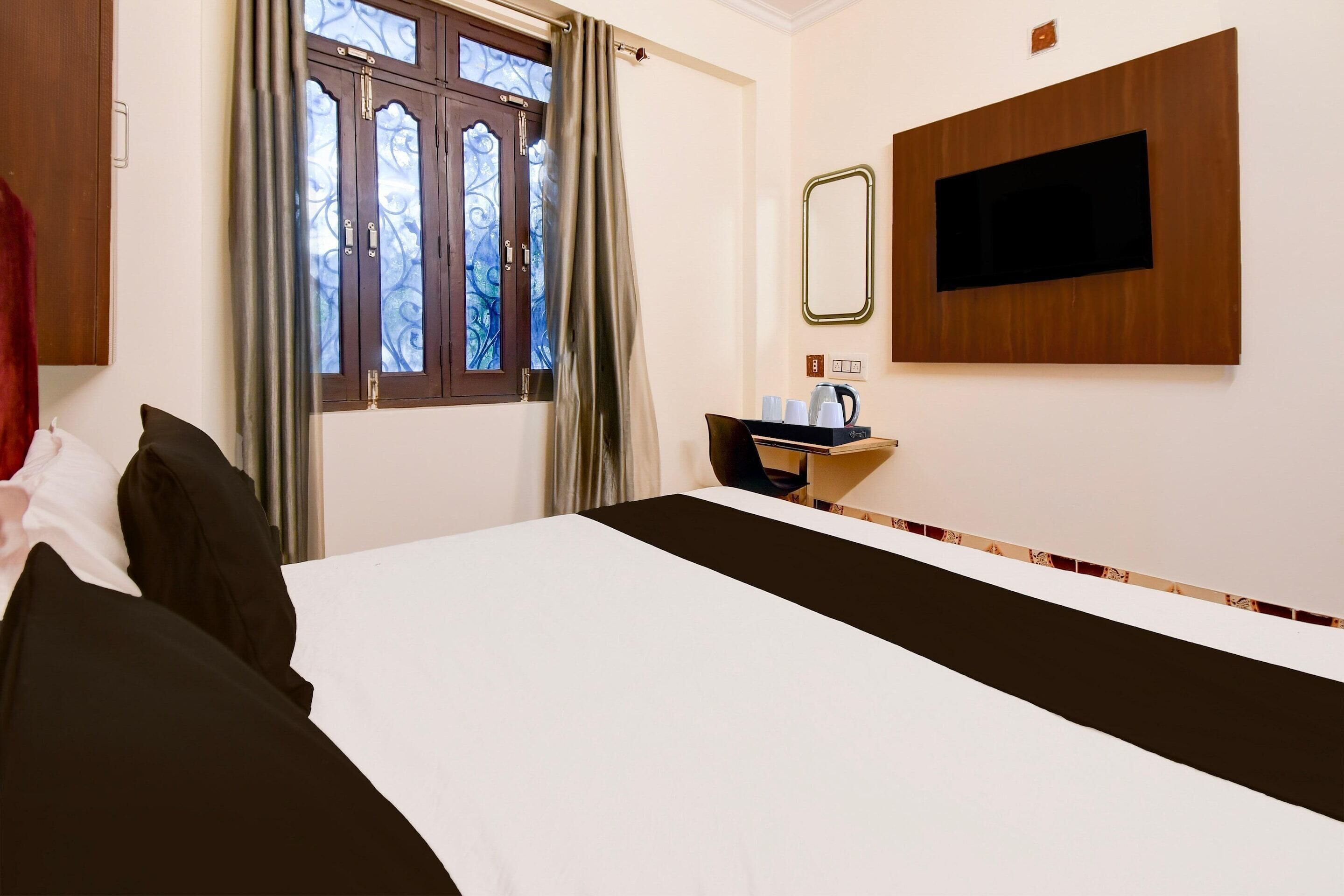 A hotel room with a bed, a TV mounted on the wall, a window with curtains, and a small table with a kettle.