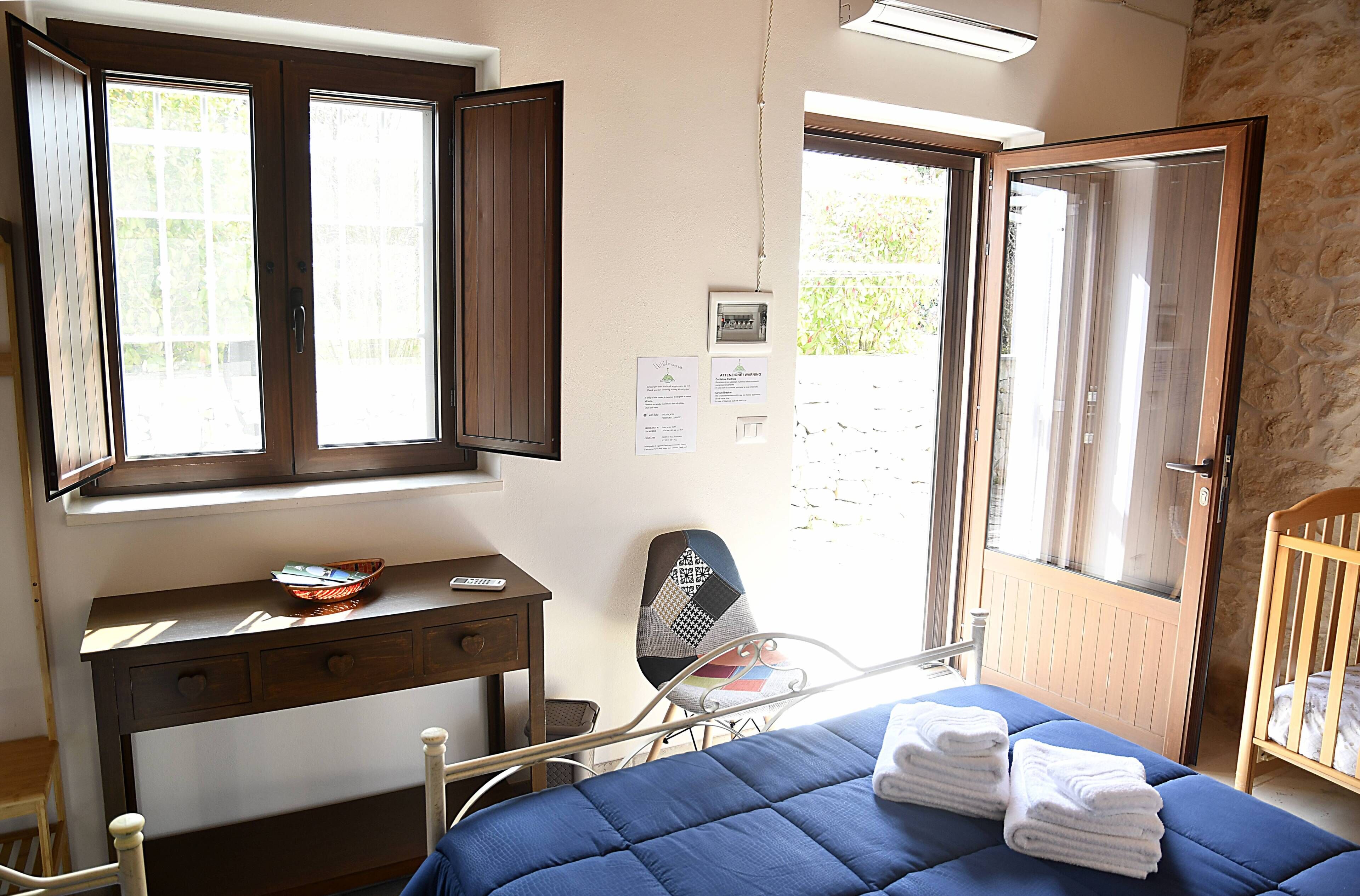 A bedroom with a bed, a desk with drawers, a chair, a crib, and a window with open shutters.