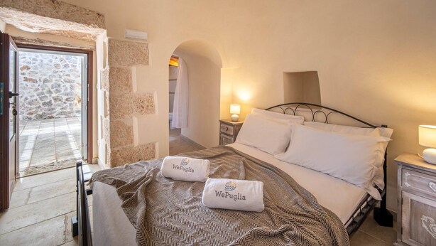 Habitación de hotel con pared de piedra, cama con colcha estampada, dos toallas con la marca 'WePuglia', una mesita de noche con lámpara y vistas a un patio de piedra.