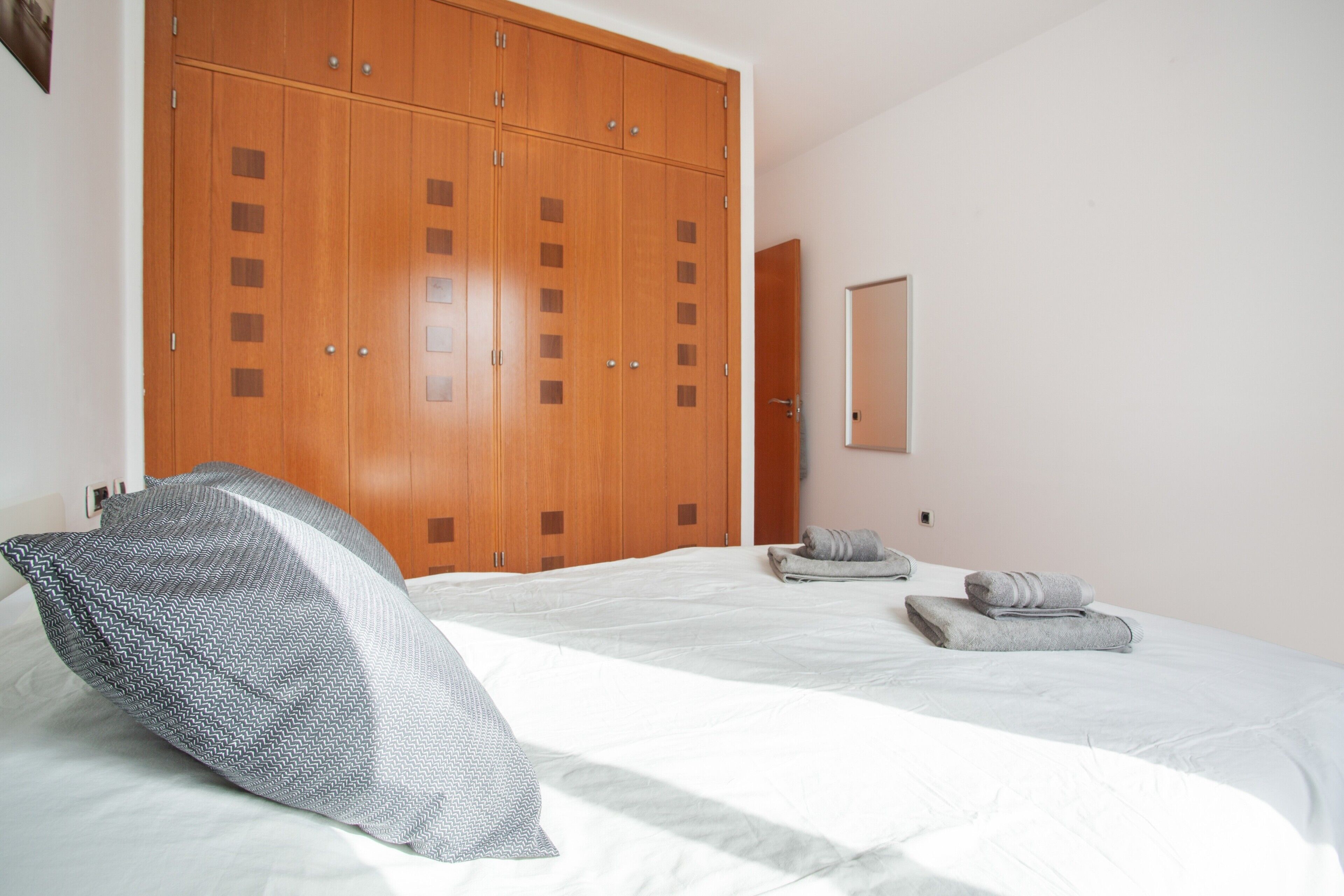 A bedroom with a large wardrobe, a bed with a patterned pillow, and two rolled towels on the bed.