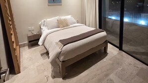 A modern bedroom with a large bed, a nightstand, a wall-mounted artwork, and a large window with a view of the city at night. - Sleek New Condo with Island Charm in Cozumel (Cozumel)