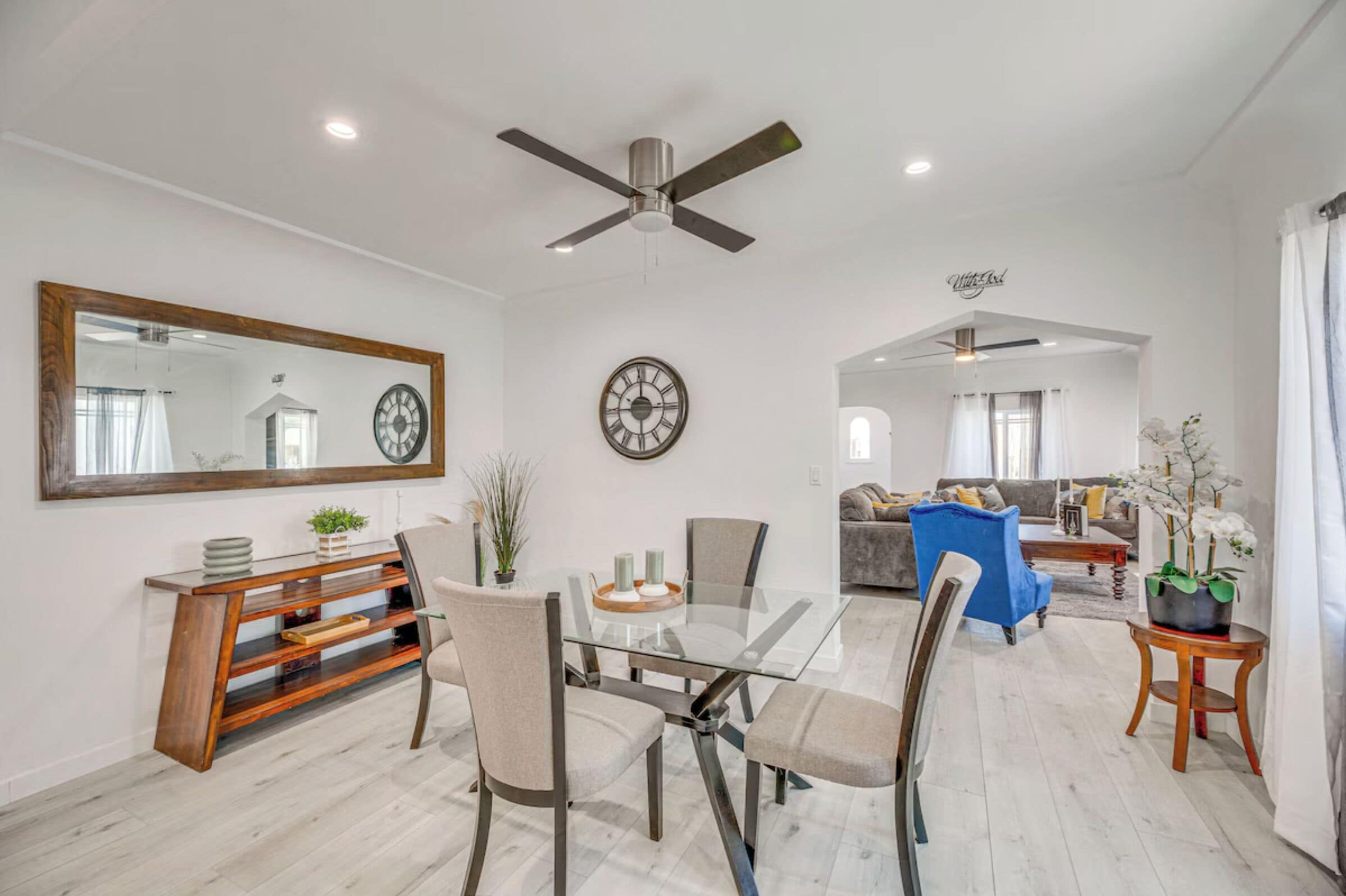 A modern living room with a glass dining table, a ceiling fan, a clock, and a sofa.
