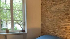 Un dormitori amb un llit, una finestra amb plantes, una paret de pedra i un paviment de fusta.