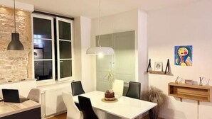 Una zona de menjador moderna amb taula blanca, cadires negres, una làmpada penjant i vista a la cuina a través d'una finestra.