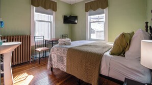 A bedroom with a bed, a desk with a chair, a radiator, and a TV on the wall. - Old Clark Inn (Marlinton)