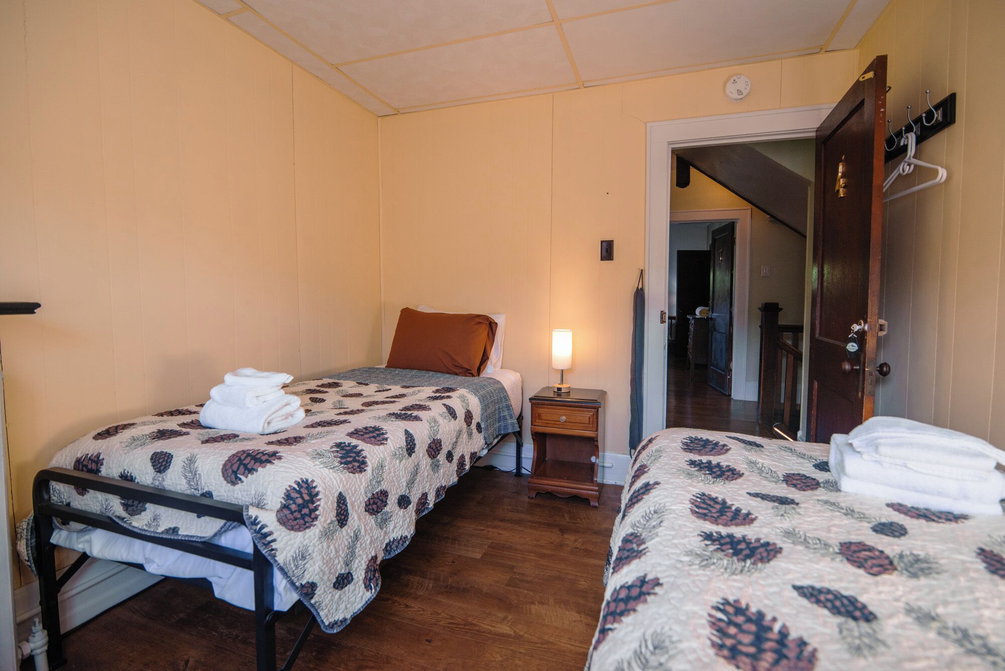 Una habitación con dos camas, una mesita de noche con lámpara, una puerta de madera y un pasillo que conduce a otra habitación.