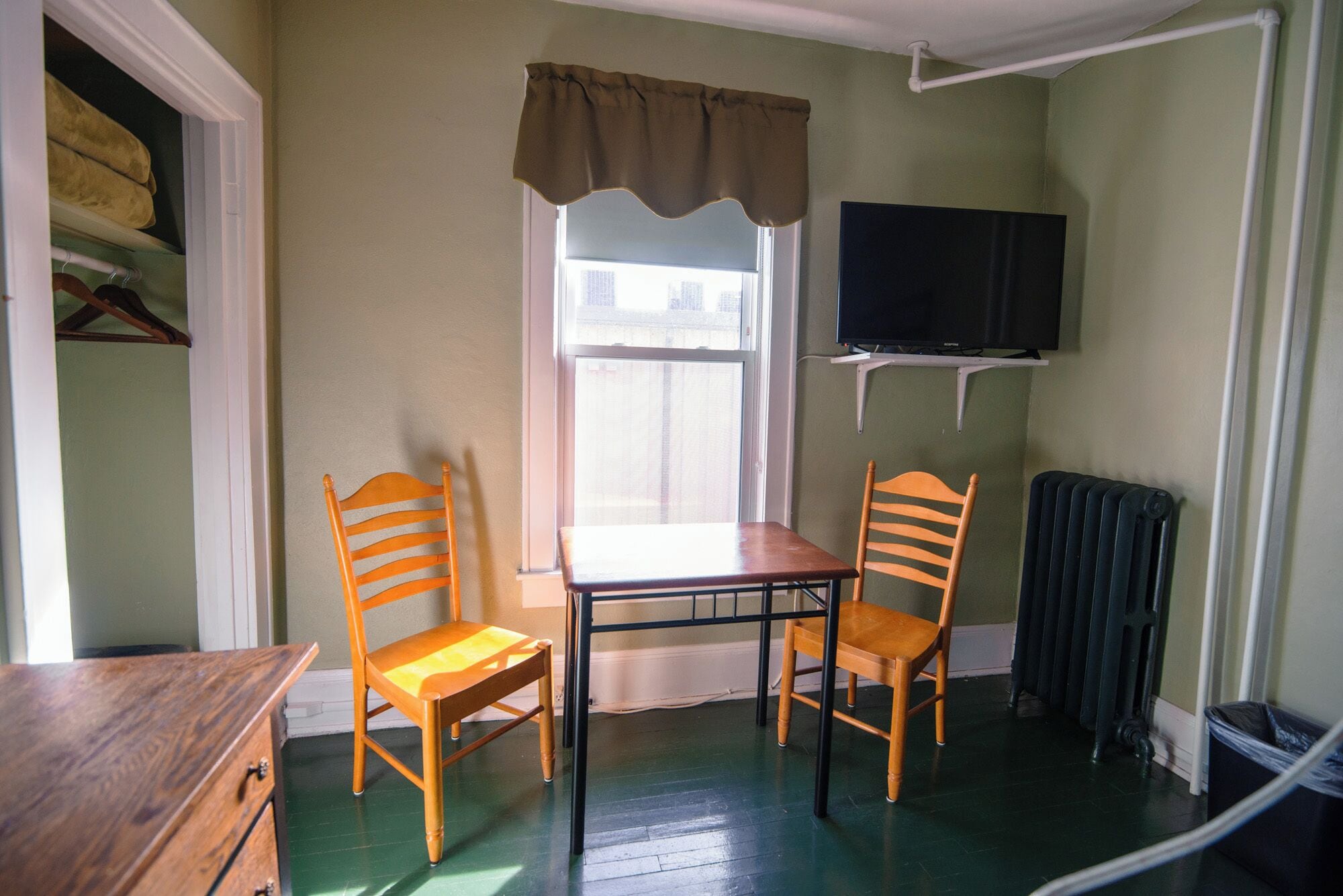 A room with two wooden chairs, a table, a window with a curtain, a radiator, and a television on the wall.