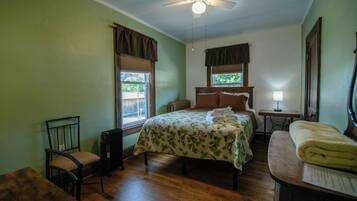A bedroom with a bed, a chair, a table, a ceiling fan, and a window.