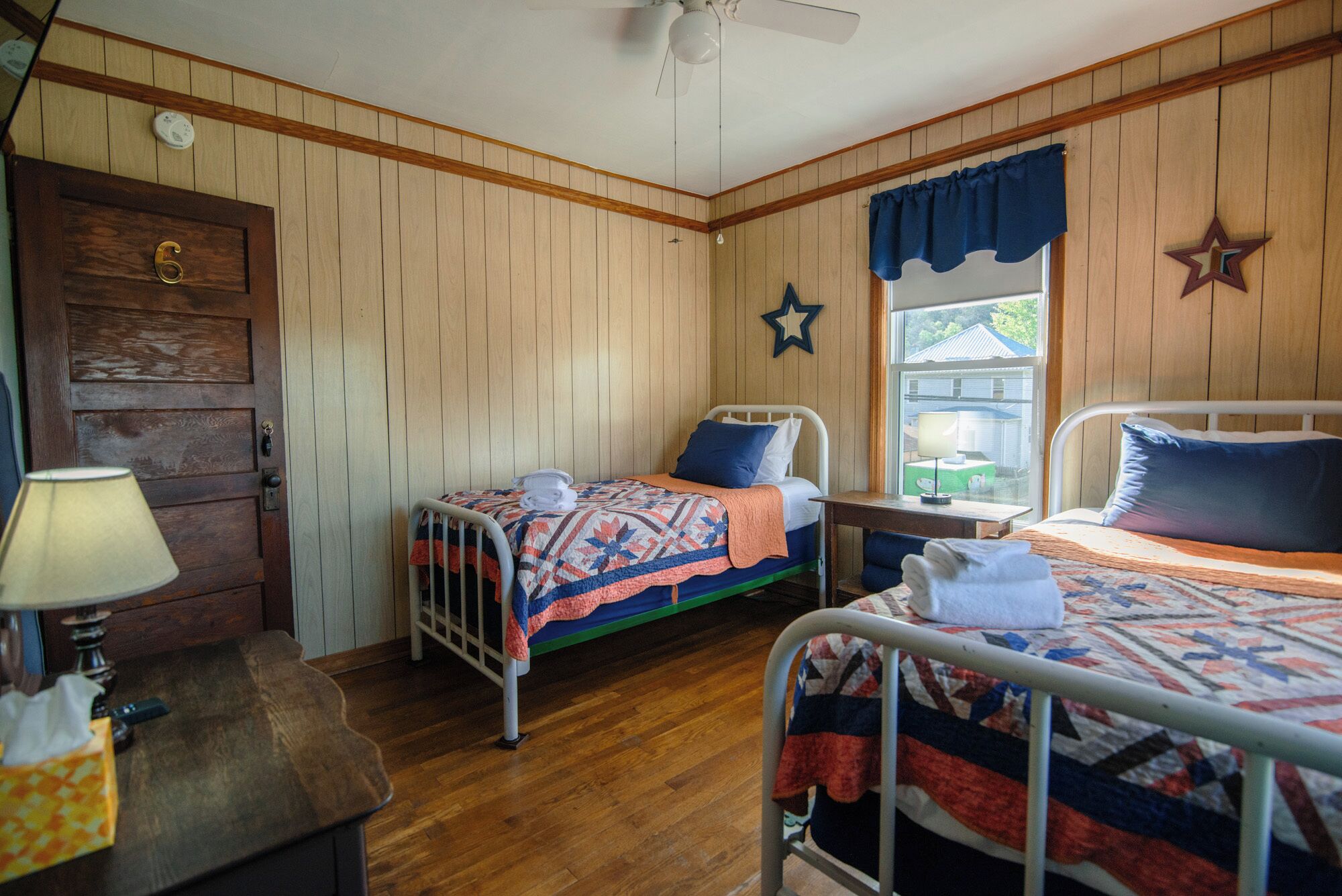 A room with two beds, wooden walls, a door, a window with a star decoration, and a ceiling fan.