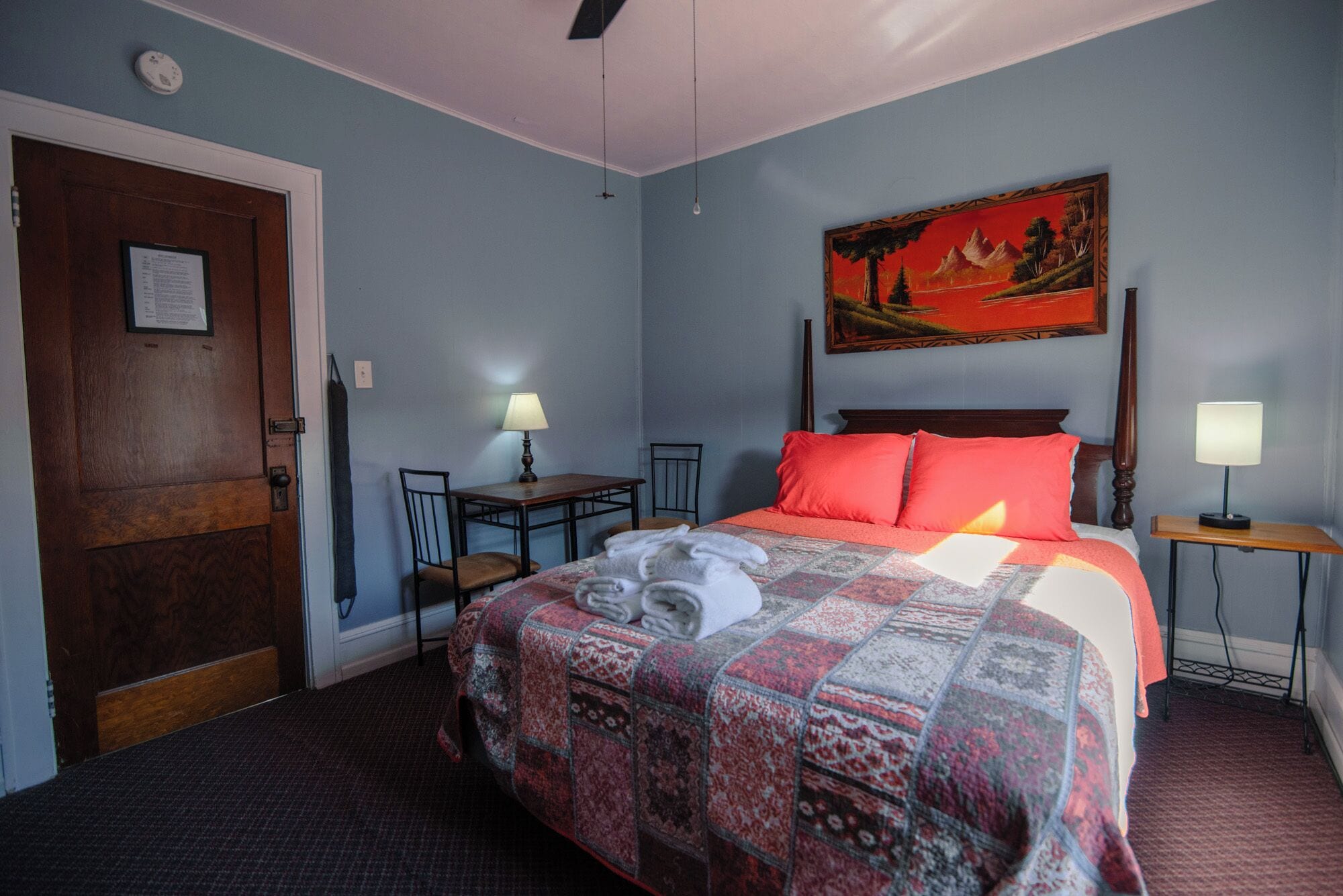 A hotel room with a bed, a desk, a chair, a painting on the wall, and a door.