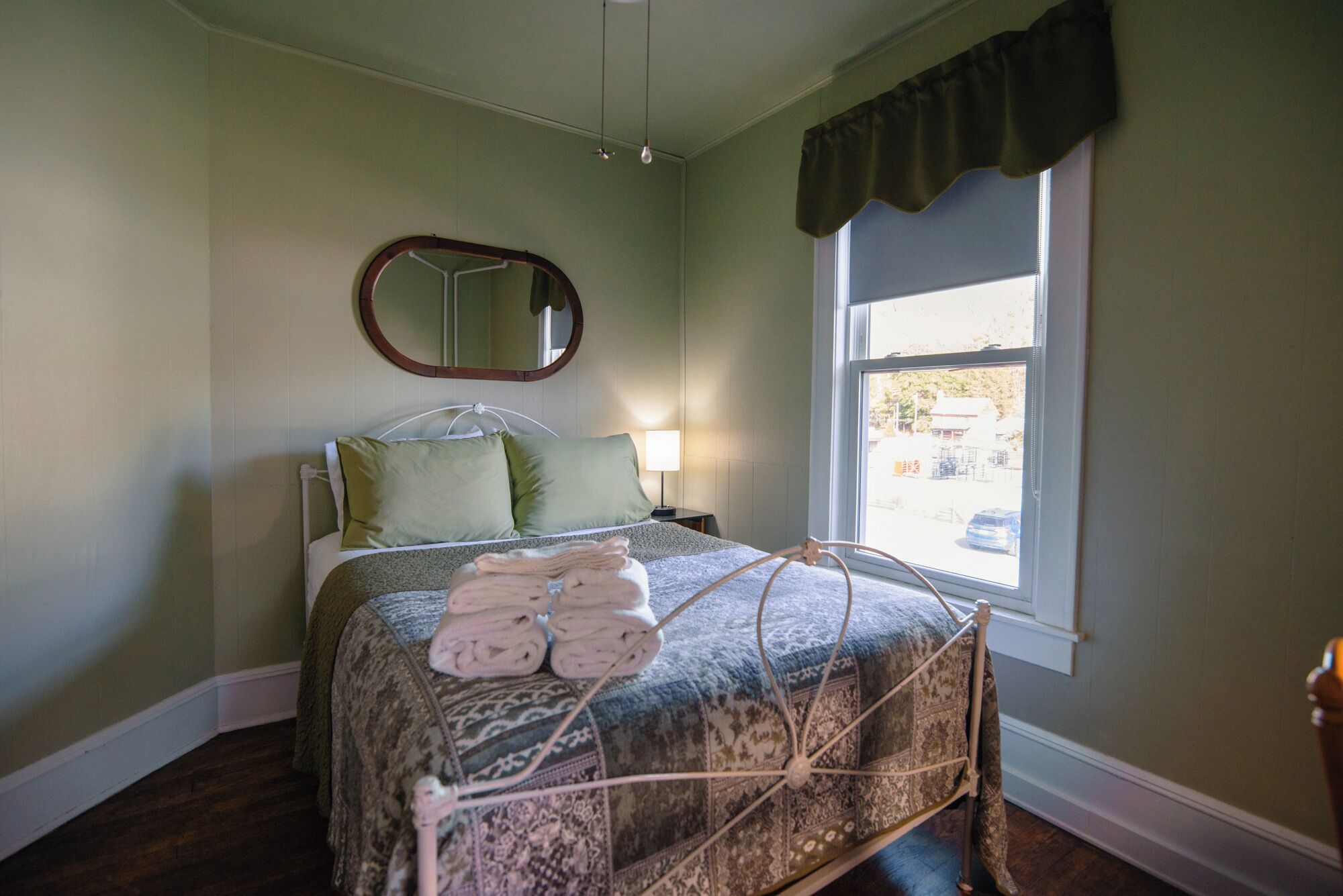 A bedroom with a bed, a bedside lamp, a window, and a mirror.