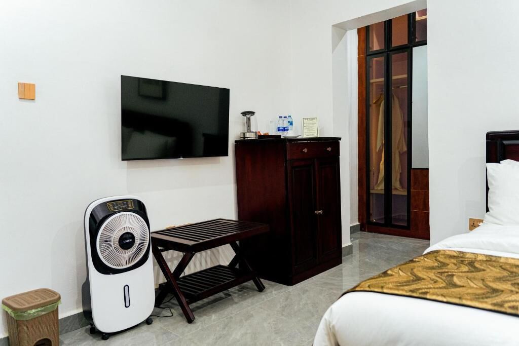 Habitación de hotel con cama, televisor montado en la pared, escritorio con silla, armario y ventilador de pie.