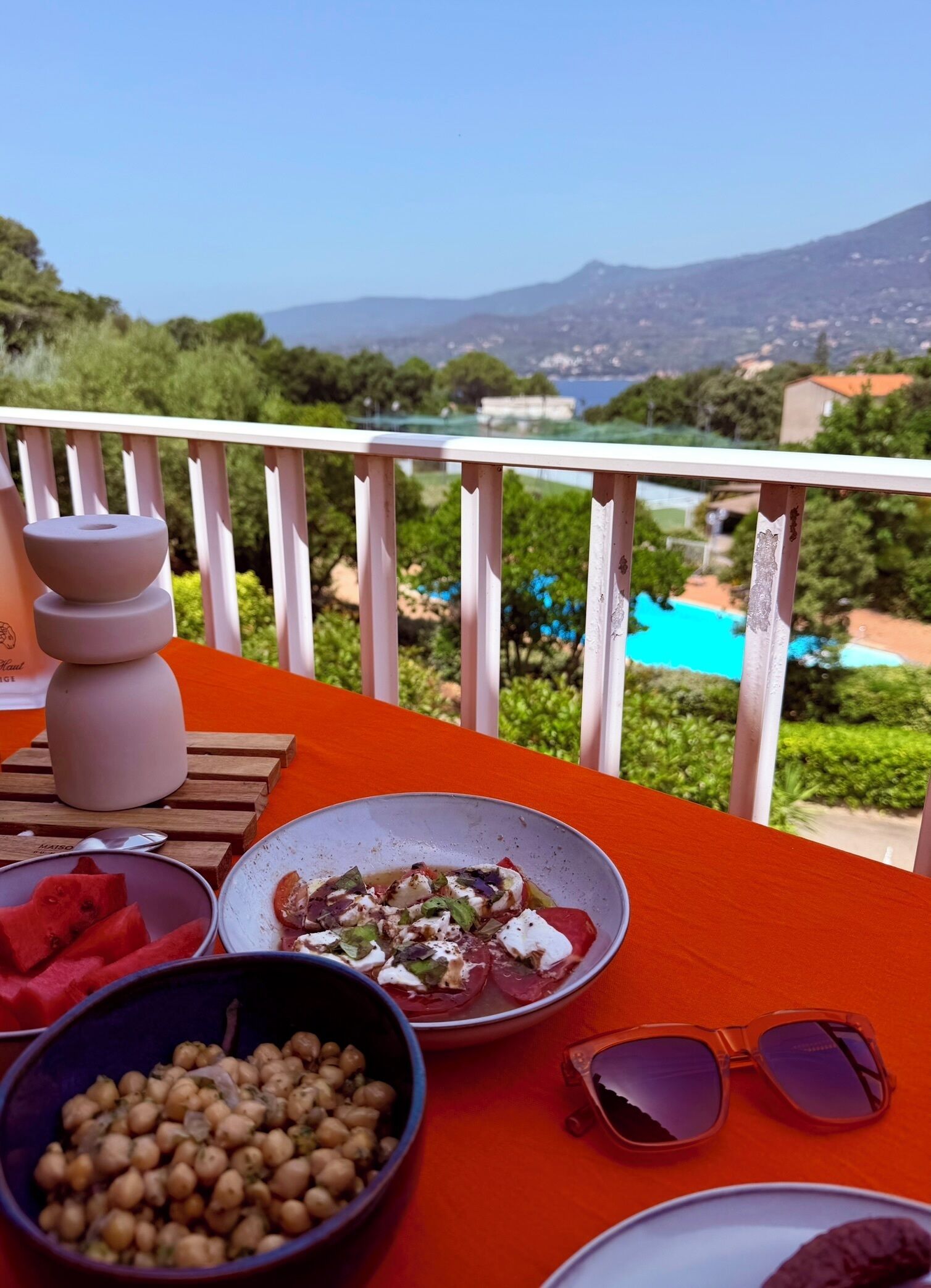 A balcony with a table set for a meal, including bowls of food, a pair of sunglasses, and a candle.
