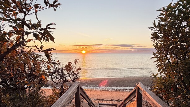 Una scala di legno conduce a una spiaggia con il tramonto sullo sfondo.
