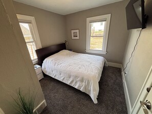 Ein Schlafzimmer mit einem Bett, zwei Fenstern, einem Nachttisch, einem an der Wand montierten Fernseher und einer Tür.