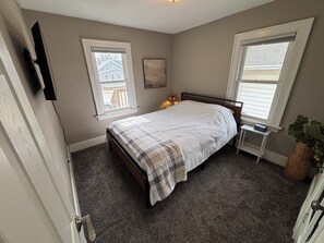 A bedroom with a bed, two windows, a bedside table with a lamp, and a wall-mounted TV.