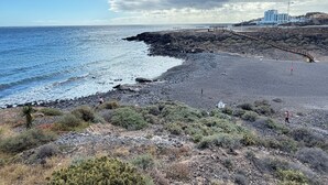 Uma praia costeira com terreno rochoso, um deque de madeira e um edifício ao fundo.