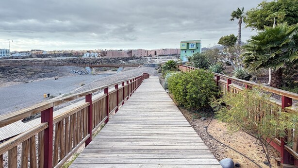 Um calçadão de madeira que leva a uma praia, com uma palmeira e um pequeno edifício ao longe.
