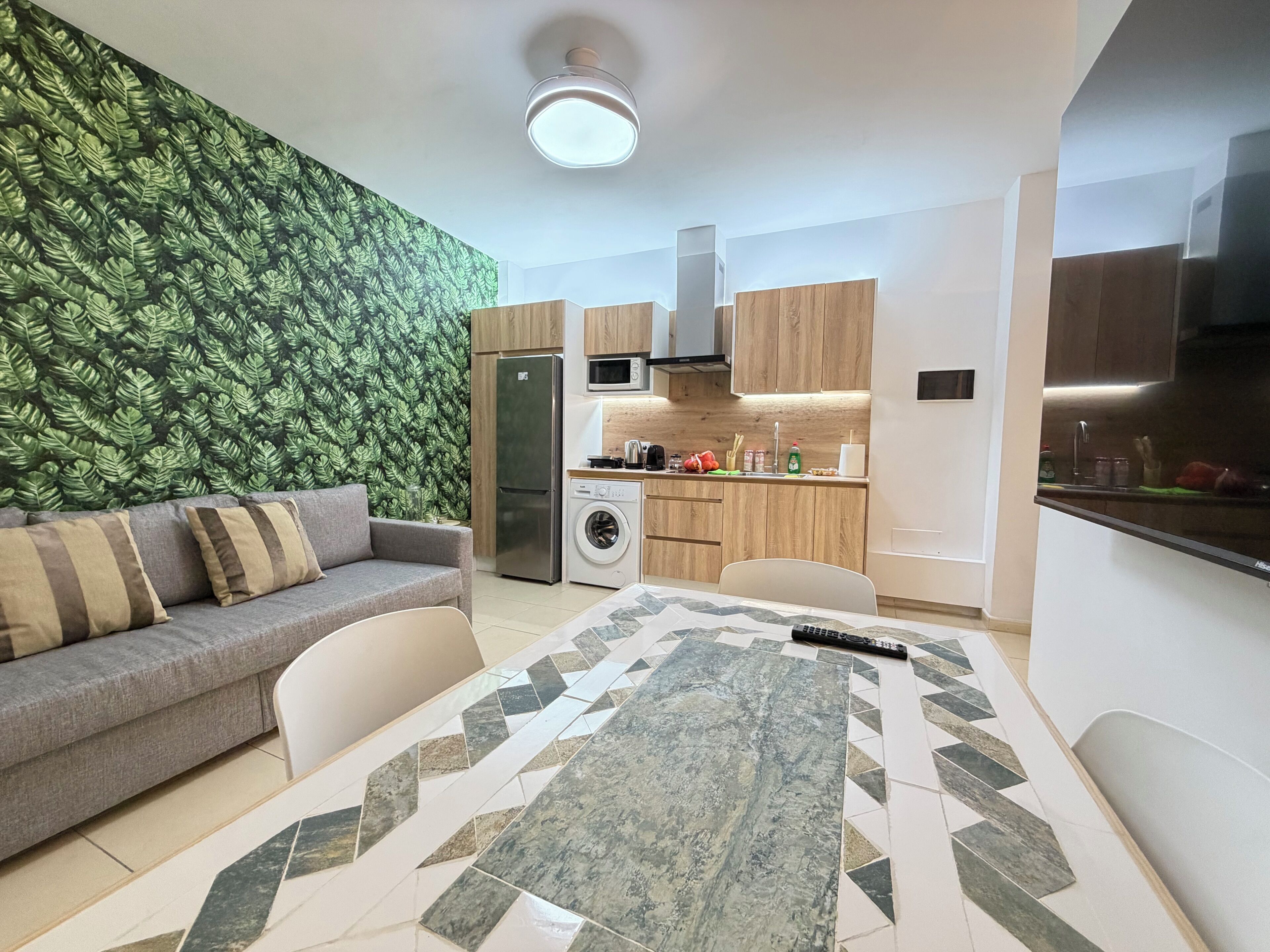 A compact living space with a sofa, a dining table, a washing machine, and a kitchen area with wooden cabinets and a microwave.