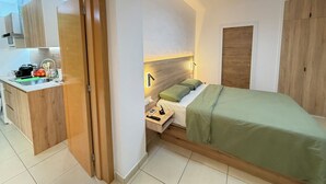 Quarto compacto com cama, mesa de cabeceira e pequena área de trabalho com luminária.