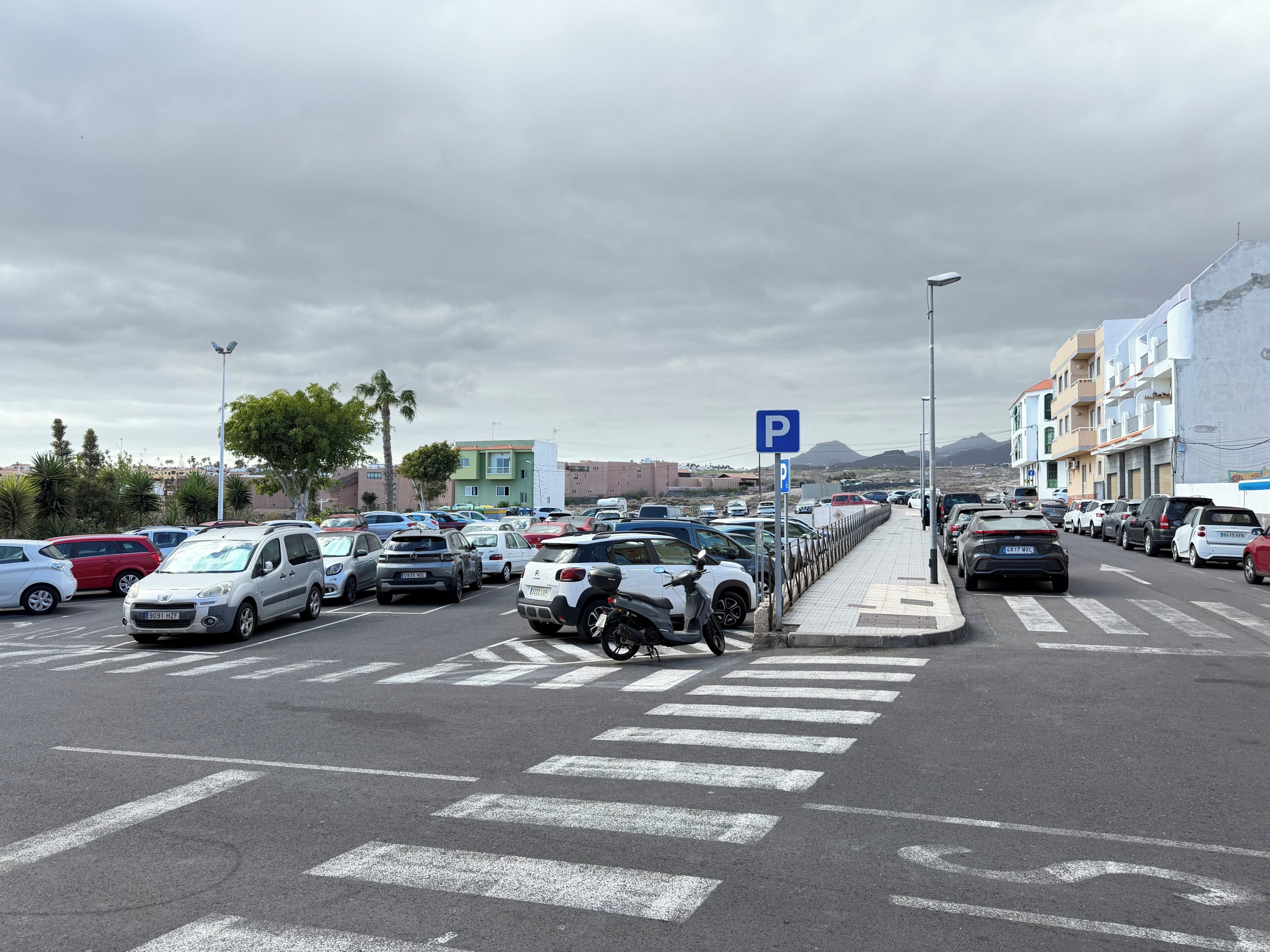 A parking lot with cars, a pedestrian crossing, and buildings in the background.