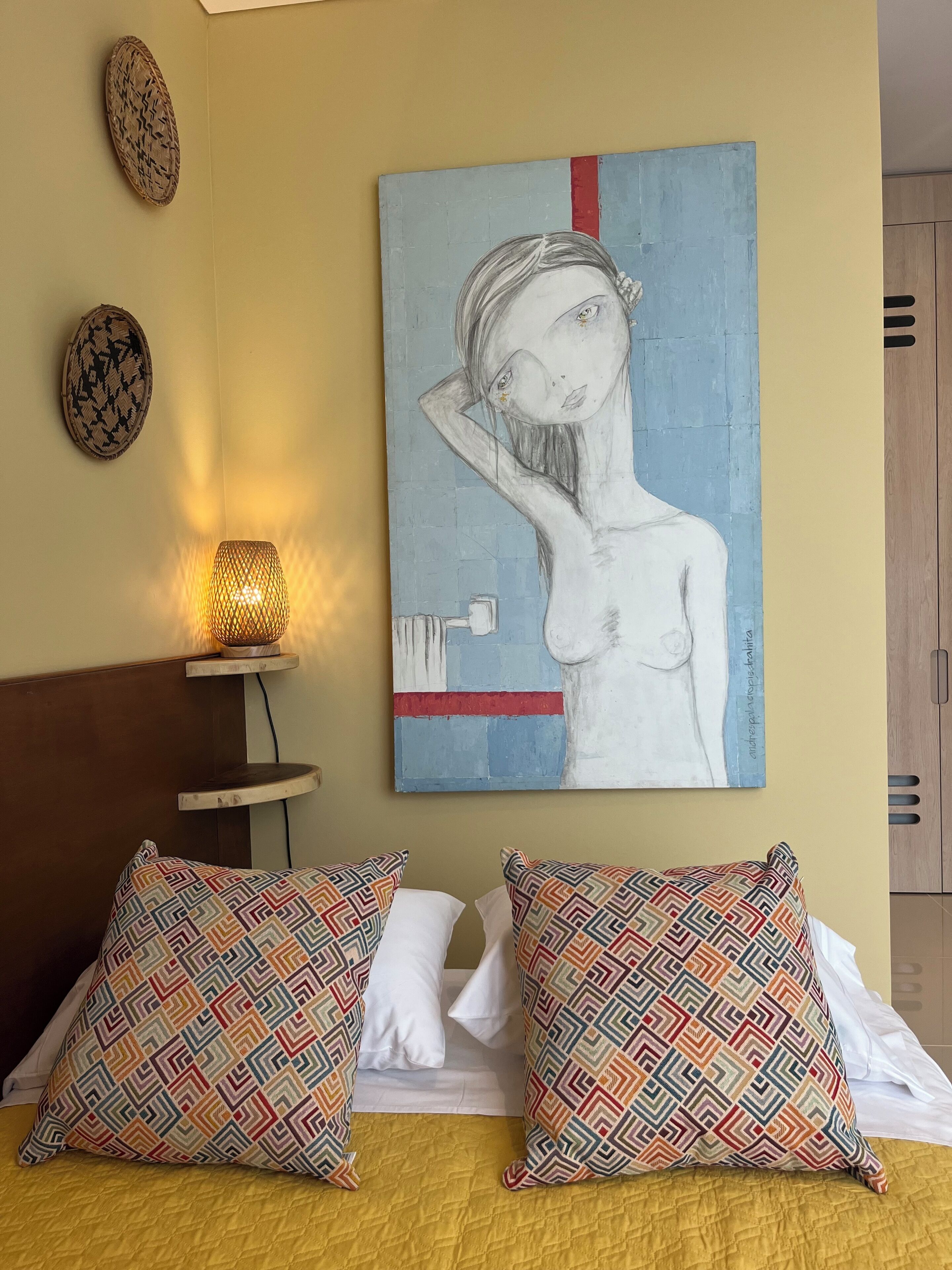Une chambre à coucher avec un lit, des coussins à motifs, un tableau accroché au mur, une petite table avec une lampe et une armoire.