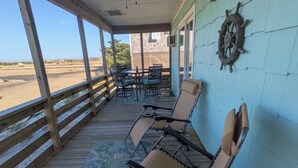 A covered outdoor seating area with a ceiling fan, a table with chairs, a decorative wall piece, and a view of the beach.