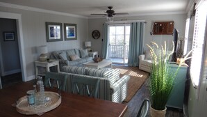 A dining area with a wooden table, chairs, and a buffet tray. There is a sofa set, a ceiling fan, and a view through the window.