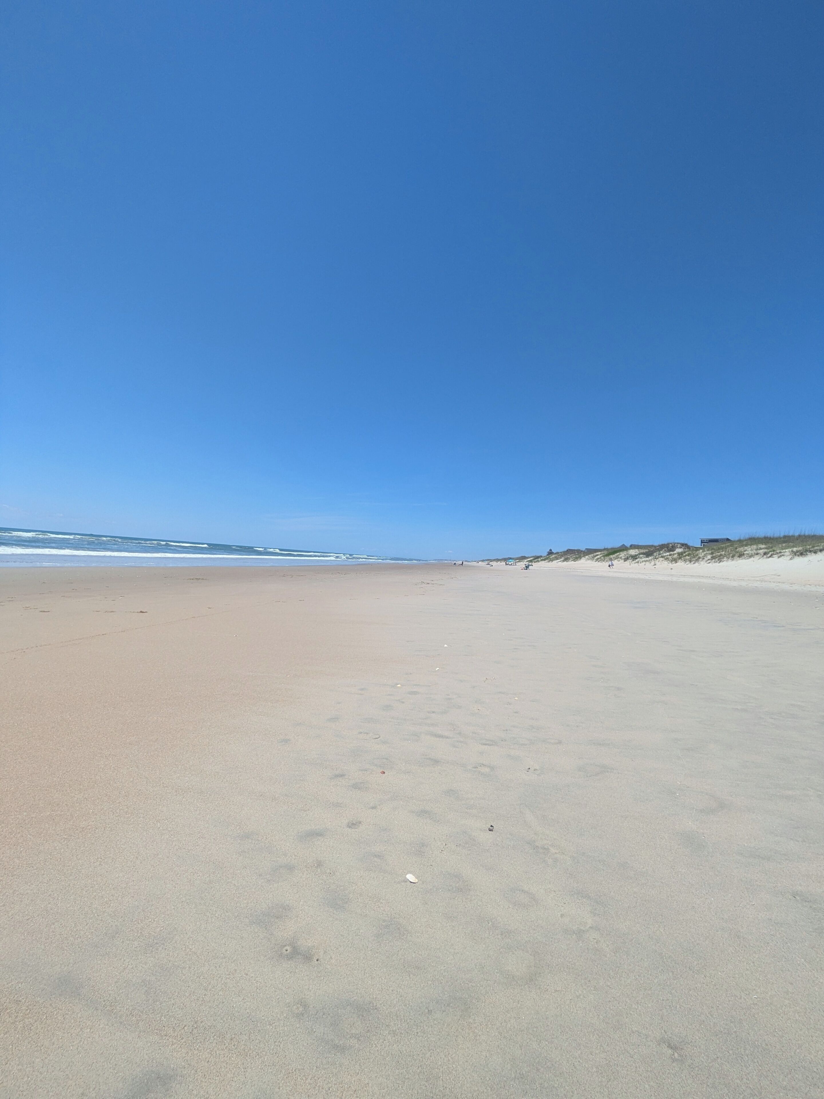 Ein weiter Sandstrand mit sanften Wellen, einem klaren blauen Himmel und fernen Dünen.