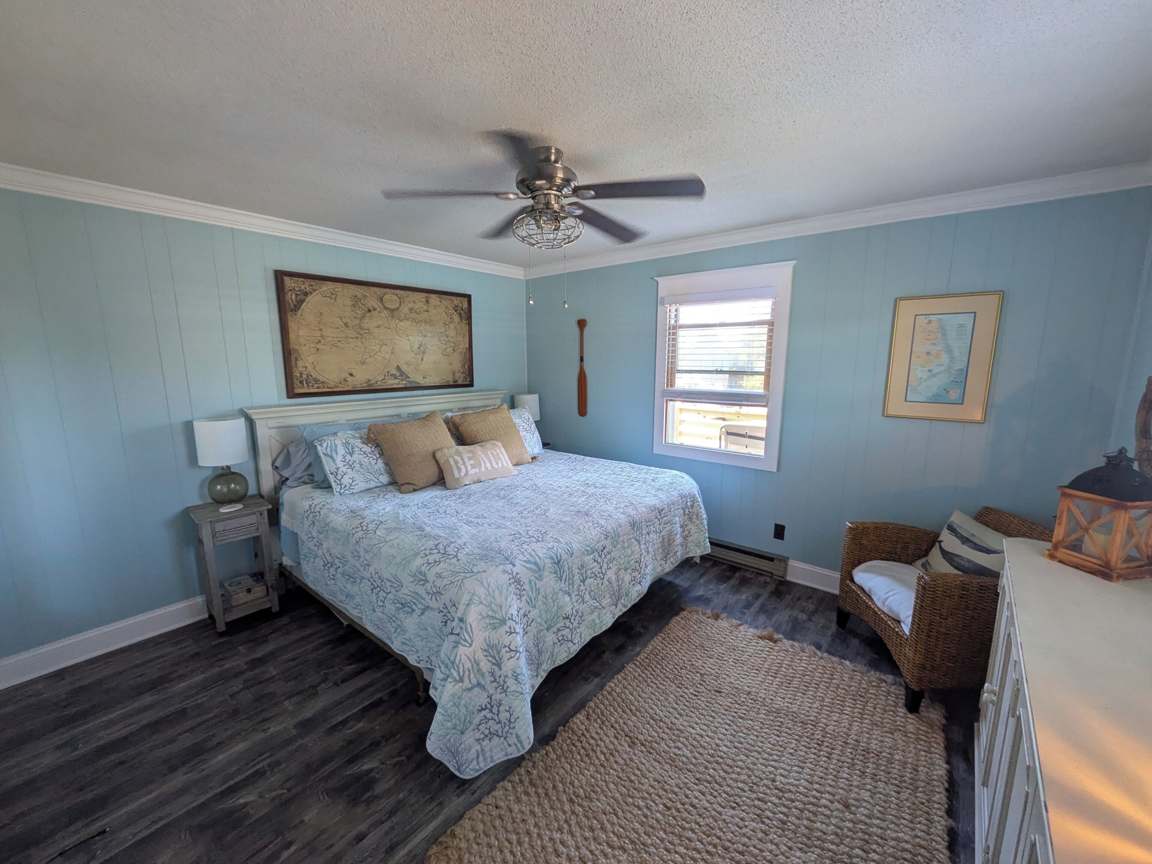A bedroom with a bed, a ceiling fan, a window, a map on the wall, and a wicker chair.