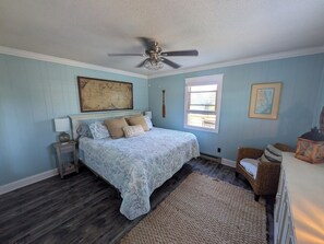Un dormitorio con una cama, ventilador de techo, ventana, mapa en la pared y una silla de mimbre.